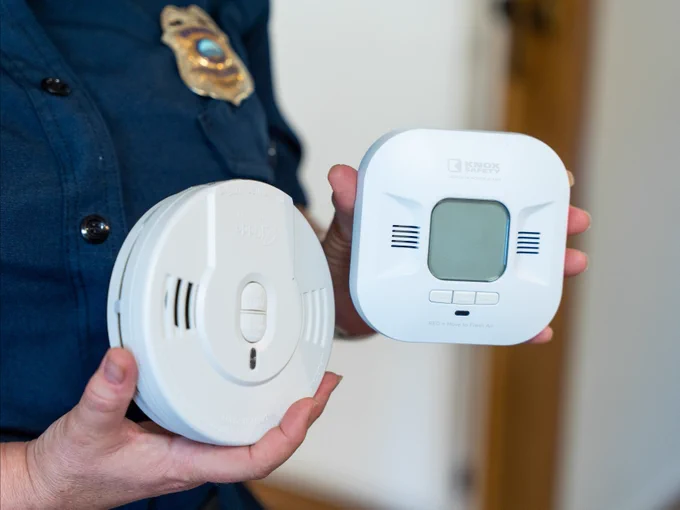 Close up photo of Huntington Beach Fire Marshal Janice Van Mullem holding a smoke alarm in one hand and a carbon monoxide detector in the other during a home safety education visit. The detectors are displayed in front of her fire department uniform to demonstrate the importance of working smoke and CO alarms in the home.