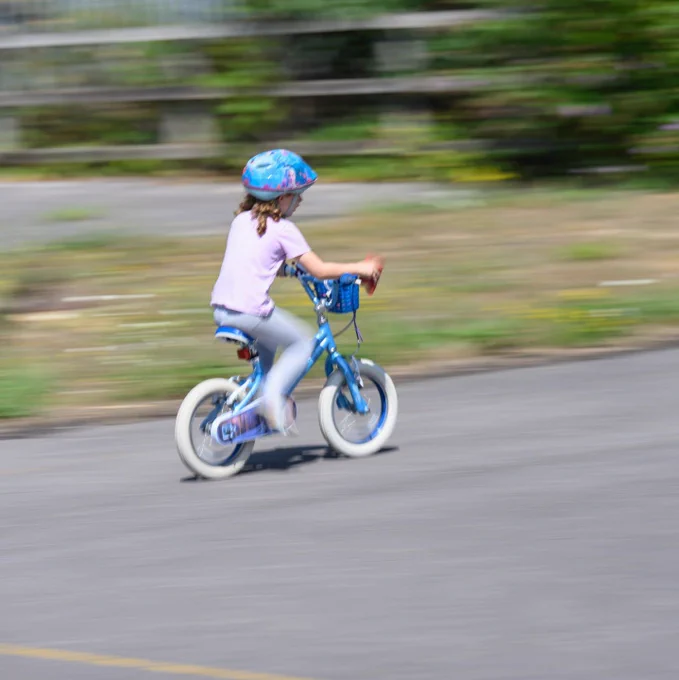 A girl on a bike pedalling away from the camera on a small child's blue bike, it looks like she is cycling very fast as all the background is a blur. 
