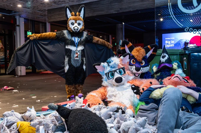 Blorange and a purple fox both sit in a rat plushie pool cuddling while there is a bat suiter behind them doing a T-pose