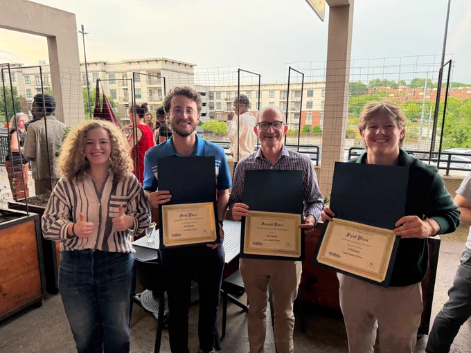 A group of four people stand on a rooftop patio. Three hold certificates, smiling proudly. The mood is joyful with a cityscape in the background.