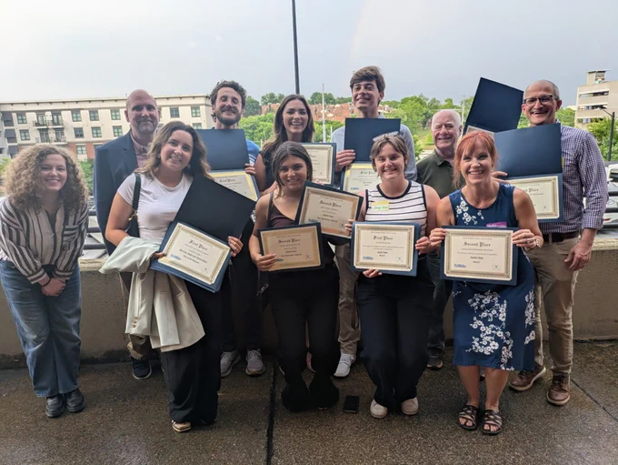 A group of smiling people stand outdoors holding certificates, celebrating an achievement. A rainbow is faintly visible in the cloudy sky behind them.