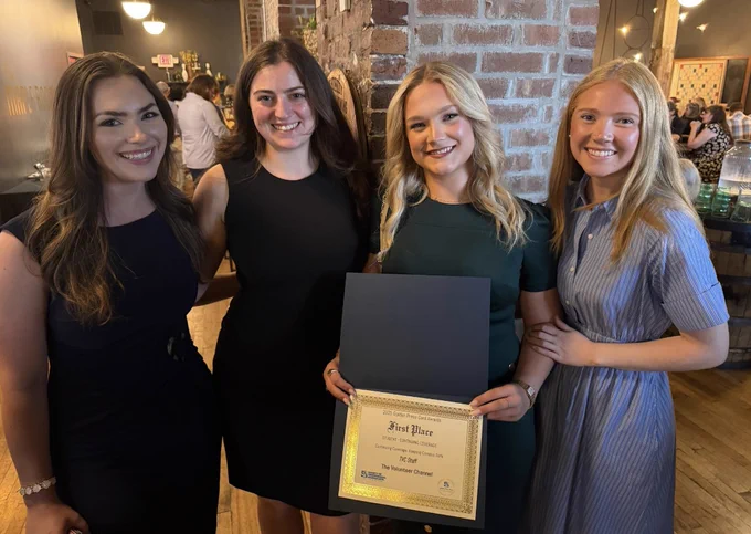 Four women smile at a celebratory event in a warm, inviting setting. One holds a "First Place" certificate. The mood is joyful and proud.