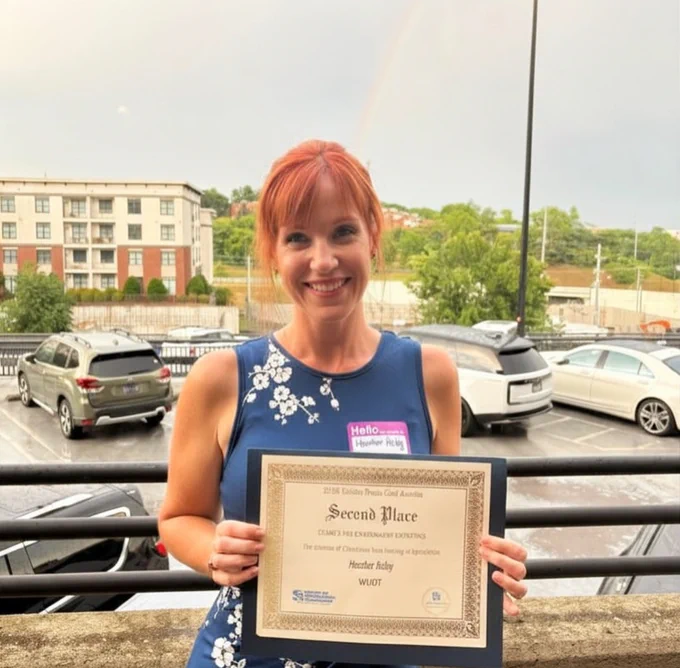 A smiling woman with red hair, wearing a blue dress, holds a second place certificate. She stands outside, with parked cars and greenery in the background.