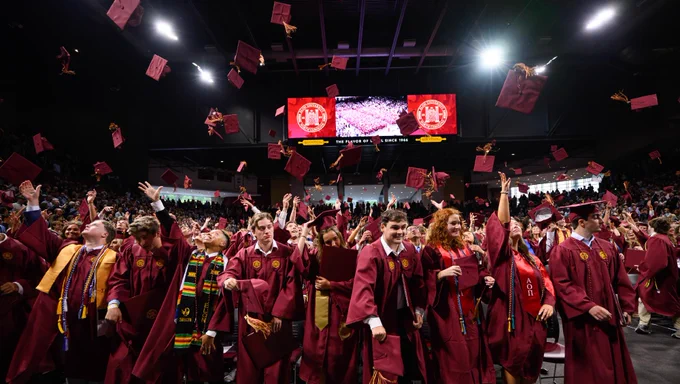 Graduates in maroon gowns celebrate by tossing caps into the air at a commencement ceremony in a large indoor venue. A screen in the background displays a university emblem and a message congratulating the class. Family and friends are seated around the graduates.
