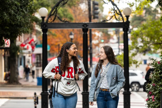 Two smiling students walk together on a university campus, passing in front of The Arch. The student on the left wears a white "Georgia Bulldogs" sweatshirt and a backpack, while the student on the right wears a blue patterned shirt over a striped tee with a small "G" logo. In the blurred background, autumn trees and a city street are visible.
