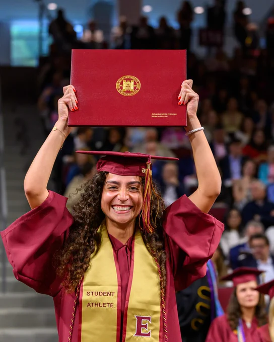 A graduate in a maroon cap and gown holds up a diploma with a gold seal. The person wears a sash labeled "Student Athlete" with the letter "E." Blurred audience in the background.