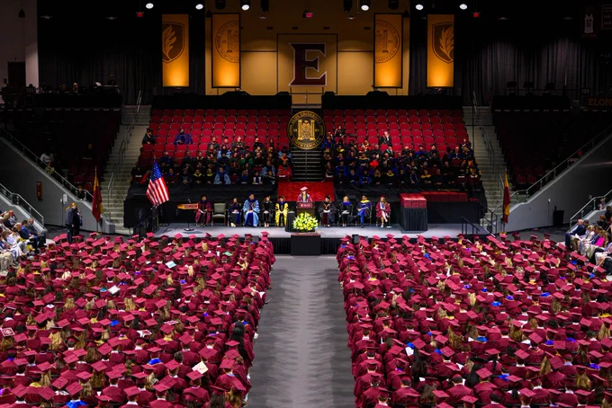 A large indoor graduation ceremony at Elon University with graduates in maroon caps and gowns seated in rows facing a stage. The stage features university officials, flags, and a podium. The backdrop displays Elon University emblems and banners.