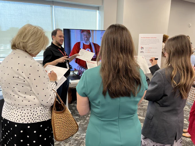 Three people stand with their back to the camera, facing a Red Cross volunteer and a TV screen. The volunteer is walking them through creating a fire escape plan on dry-erase boards.