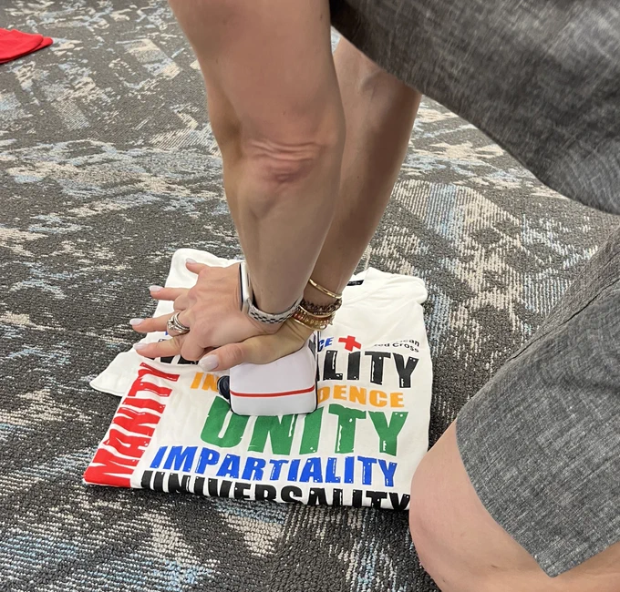 A close up of someone's hands as they practice hands-only CPR compressions on a stress toy placed on a folded t-shirt on the floor.