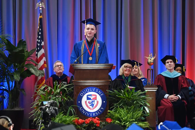 Joshua Stewart speaks from the podium in academic regalia during Commencement.
