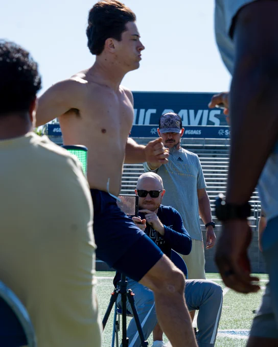 Two coaches are clocking a UC Davis student athlete with stop watches as he passes the finish line for a 40-yard dash while wearing blue shorts and no shirt