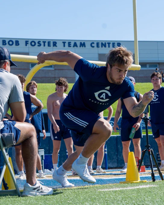 UC Davis student athlete taking off from the goal line to run a 40 yard dash while on the football field and wearing a blue shirt and blue shorts with practice players watching behind