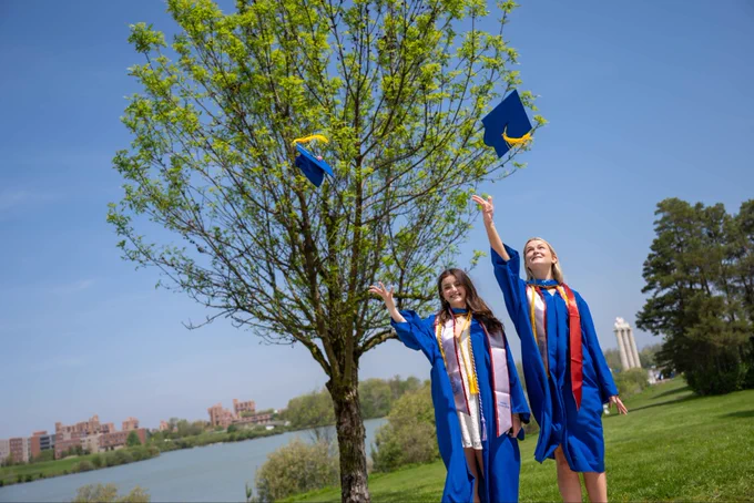 Two students toss their graduation caps in the air in front of a tree.