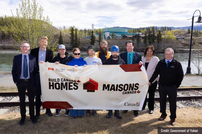 Minister Robertson and PS Hanley with a group of people holding a large banner that says “Build Canada Homes” with the river and trees behind them and the  hospital in foreground on the opposite embankment.