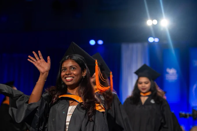 A smiling woman in a graduation gown waves, with others in similar attire behind her. They wear black gowns and caps with orange tassels, illuminated by bright lights against a dark blue backdrop.