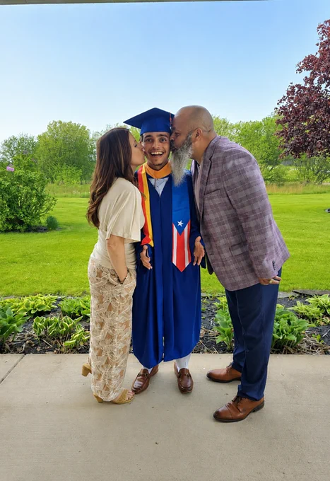 A graduate is being kissed on the cheeks by his mother and father. The graduate is wearing a blue graduation gown and cap, and is smiling. The mother and father are smiling at @number, and trees are in the background.