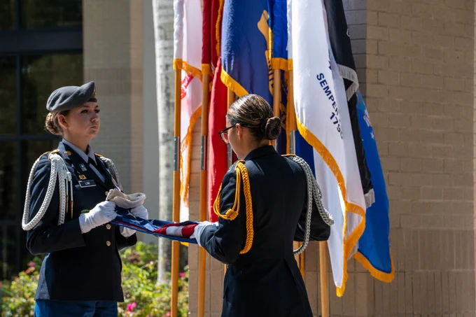 Two uniformed individuals participate in a flag-folding ceremony. One person holds the folded American flag while the other assists. They are in formal military attire, wearing gloves and decorative cords. Behind them, several flags are displayed.