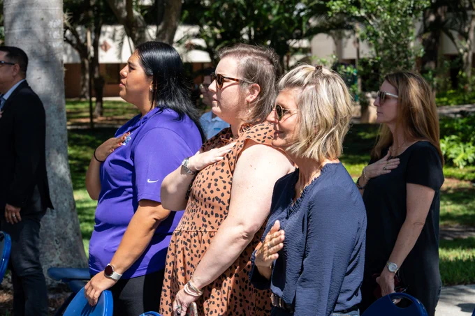 People stand outdoors with their right hands over their hearts, appearing to participate in a ceremonial or patriotic event. They are in a row, facing an unseen focal point, surrounded by trees and greenery. The women wear casual outfits, including a purple shirt, a leopard print dress, and sunglasses.