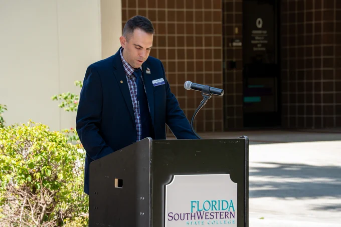A man in a dark suit, red and blue plaid shirt, and a tie is speaking at a podium with a microphone. The podium displays the sign "Florida SouthWestern State College." The background features a building with brown tiles and foliage to the left.