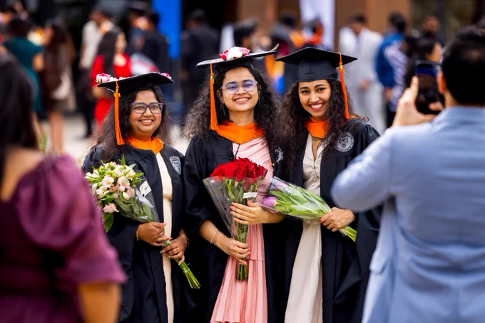Three smiling young women in graduation gowns pose with flowers; person takes their photo. They wear caps with orange tassels and academic hoods. The women appear to be of South Asian descent. 'STATE UNIVERSITY OF NEW YORK' emblem is visible on the gowns.