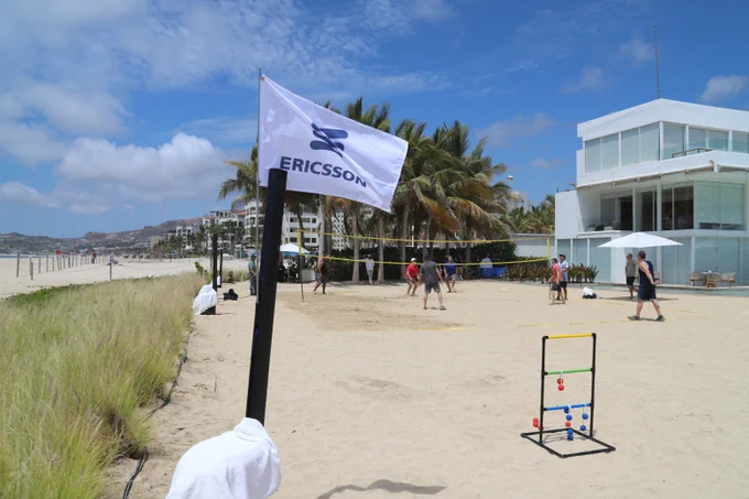 Ericsson employees play beach volleyball beside an Ericsson flag at a coastal resort during a company gathering ahead of Memorial Day weekend, recognizing time for reflection, connection, and honoring those who served.