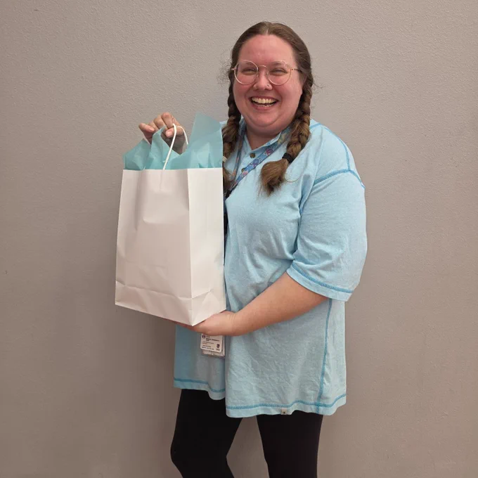 A cheerful woman in a blue shirt and glasses holding a white shopping bag and smiling at the camera.