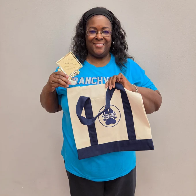 A smiling person wearing glasses, a blue shirt, and holding up a white canvas tote bag with "CANCHY" written on it.