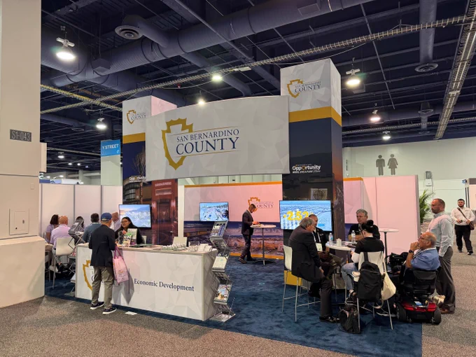 A trade show booth for San Bernardino County featuring a large banner with their logo. Several attendees are sitting and conversing at tables. There is a display with informational materials and a screen showing a presentation. 