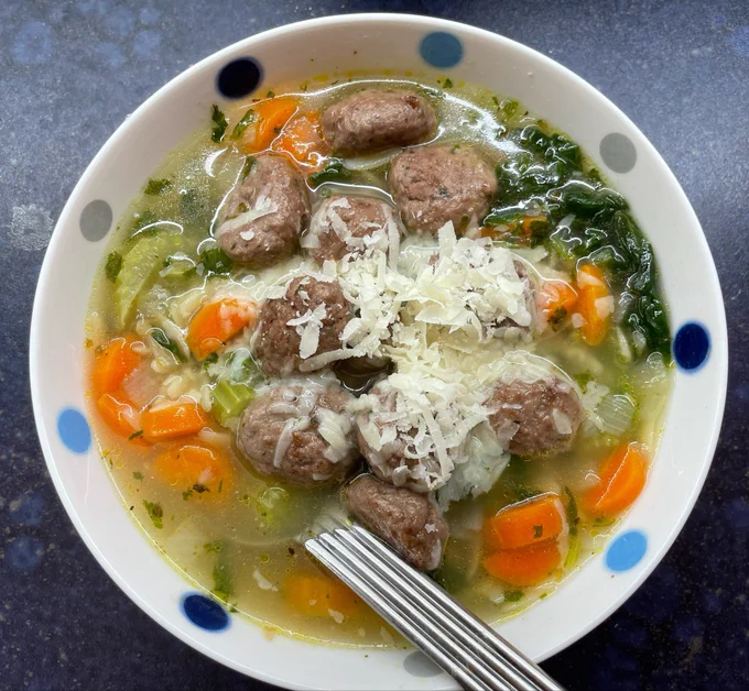 White bowl with blue spots filled with soup containing carrots, celery, onion, spinach and orzo pasta topped with meatballs scattered with Parmesan