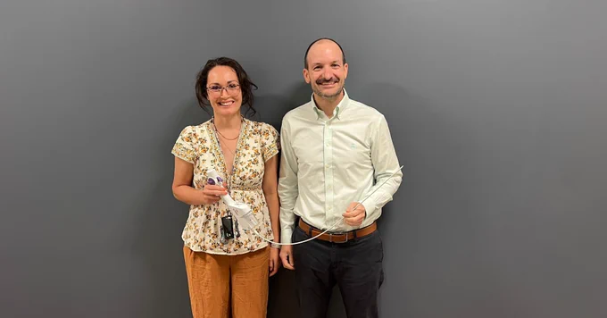 Two clinicians stand in front of a gray wall holding components of a CVAC aspiration system, including a handheld suction device and a long flexible catheter used in minimally invasive kidney stone procedures.