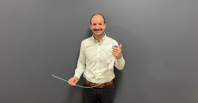 A clinician stands in front of a gray wall holding a CVAC aspiration device connected to a long flexible catheter used for minimally invasive removal of kidney stones.