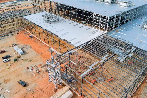 Aerial view of a large industrial building under construction. Steel framework and metal roofing dominate the site, with scattered construction vehicles and earthmoving equipment on red soil.