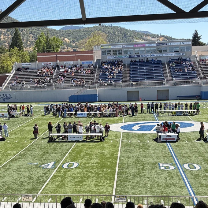 A view of a football field with several college tables set up on the field for High School Decision Day. Rogue Community College has a huge crowd of people at its table compared to the others. It's a beautiful clear sunny day.