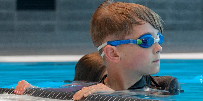 Child wearing blue goggles resting at the edge of an indoor swimming pool with tiled walls in the background.
