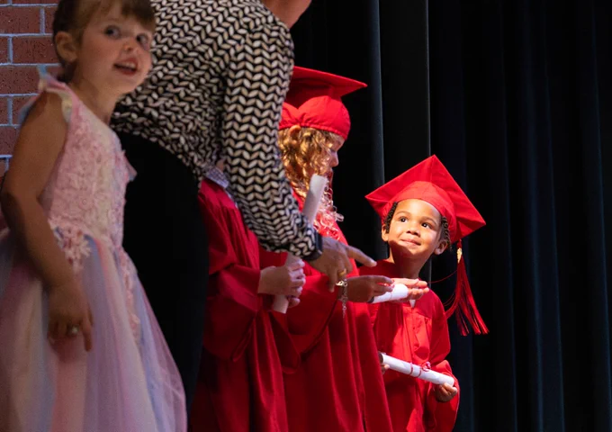 A PreK graduate stands in the spotlight smiling at her teacher as she holds her diploma.