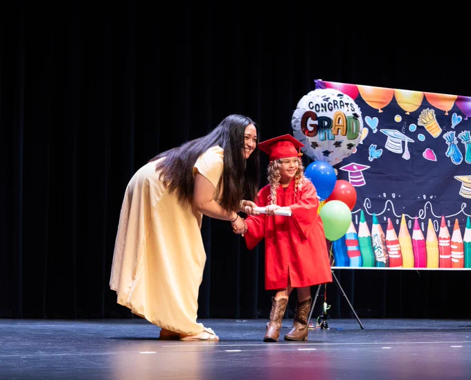 A PreK Graduate stops and poses for a photo with her teacher shaking her hand as she receives her diploma.
