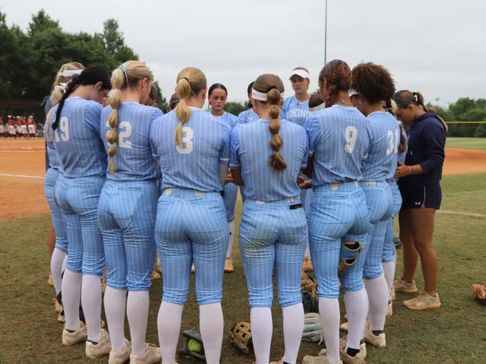Softball team in light blue pinstriped uniforms standing in a circle on the field during a team huddle.