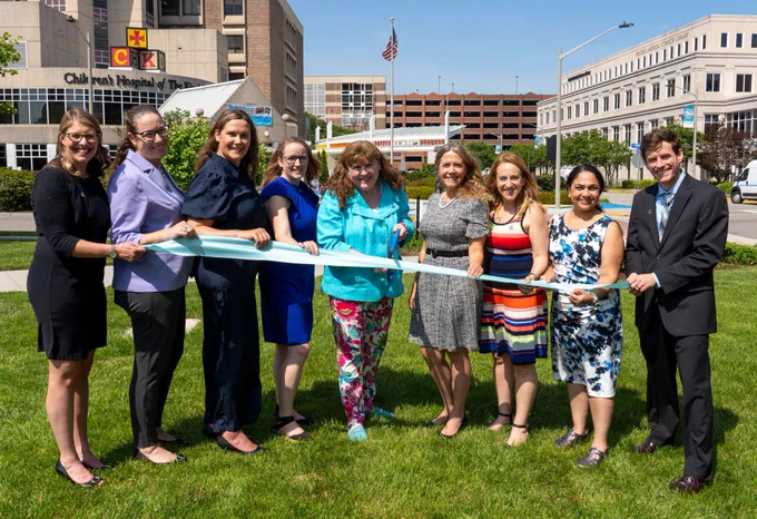 Group of nine people standing on grass outside a hospital, holding a long ribbon for a ribbon-cutting ceremony on a sunny day.