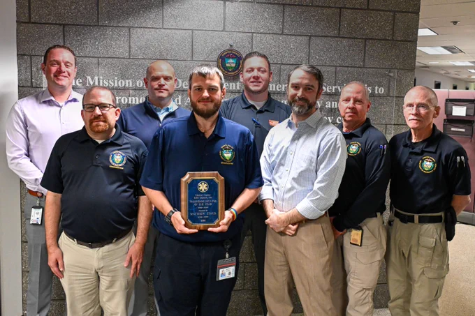 A photo of Telecommunicator Matt Staud holding his telecommunicator of the year award surrounded by a group of EMS Leadership and Department of Emergency Services leadership.
