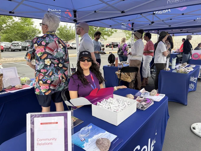 A person sits at a booth under a Select Health tent, surrounded by brochures and small items. Others are standing and interacting nearby. The setting appears to be an outdoor community event.