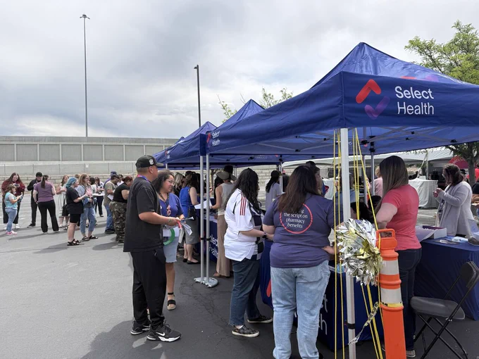 People are gathered under Select Health tents at an outdoor event. There are tables with various items, and some individuals are interacting with representatives at the tables. The setting is a paved area with trees and a cloudy sky.