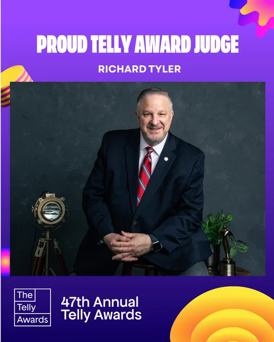 Richard Tyler, Telly Awards Judge, seated in a dark blue suit with a white shirt and red tie with light blue stripes, wearing his Commitment to Excellence lapel pin. He is smiling, hands folded in his lap, posed against a dark gray washed studio background. To the viewer’s left stands a vintage brass photography light on a tripod, and to the viewer’s right a small polished wooden table holds a green ivy plant and a gold Telly Award statuette.
