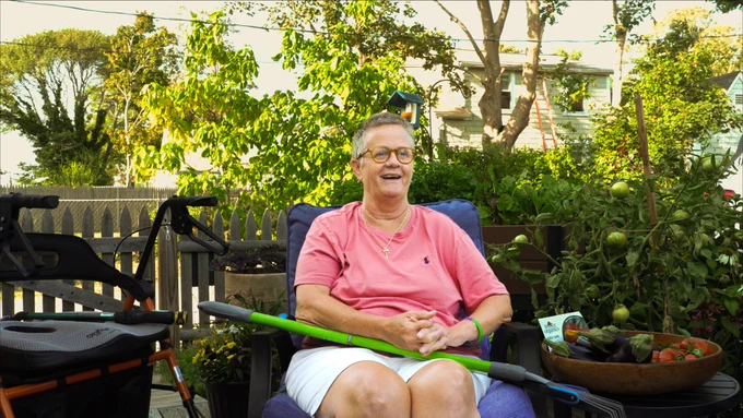 Patty Ericson-Taylor smiling while seated in a chair outside her home in her garden area. She is holding a rake and near a bowl with fresh produce inside.