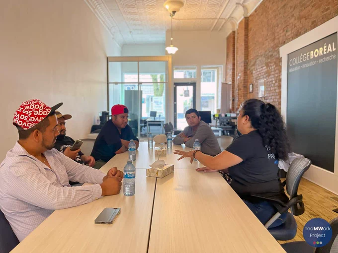 A small group of migrant workers sits around a table in a community office, listening as a service provider explains information and gestures during a support session. Bottled water and a tissue box are on the table, and a “Collège Boréal” sign is visible on the wall in the background.
