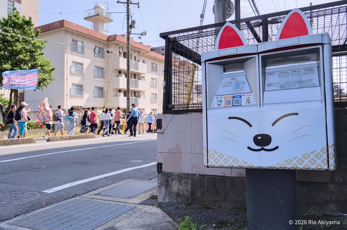 画面手前に写る、白地に黒で狐の顔がデザインされ、赤い耳がついた「きつねポスト」。その奥の車道を、レインボーフラッグやトランスジェンダーフラッグを掲げた山口レインボープライドのパレード参加者や警察官が、画面右方向に向かって歩いている。