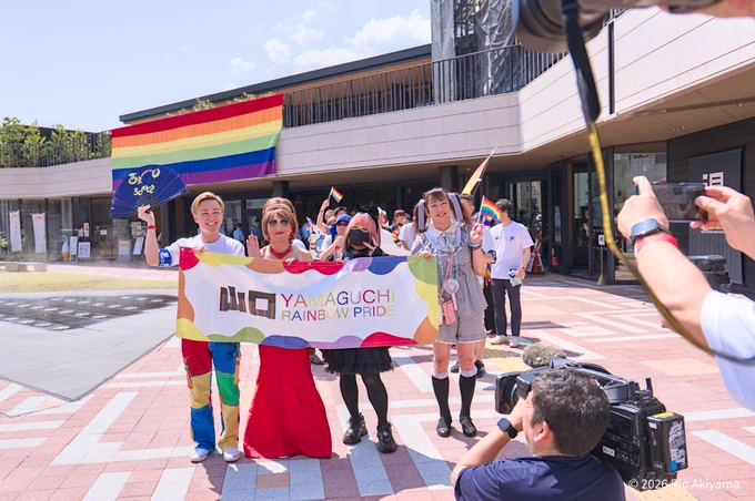湯田温泉こんこんパークの前で、「YAMAGUCHI RAINBOW PRIDE」と書かれた横断幕を広げて記念撮影をする参加者。赤いドレスのドラァグクイーンなど4人が並び、周囲ではカメラやスマートフォンを構えて撮影する人々が写っている。建物の2階部分には巨大なレインボーフラッグが掲げられている。