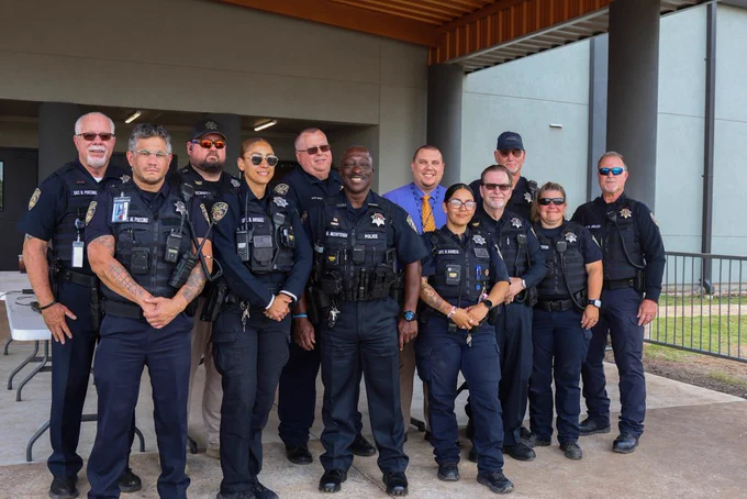 Police officers in uniform stand in a row, with two women and a man in a blue shirt. Behind them is a building with glass walls and pillars.