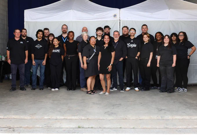 A group of people in black t-shirts and sneakers, some with lanyards, poses for a photo under white canopies.