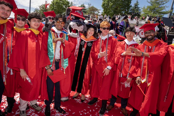 I am standing with a group of Stony Brook University students in red graduation gowns stand together with Wolfie, the Stony Brook mascot, smiling and celebrating at a commencement ceremony. Confetti is scattered on the ground, and trees and a crowd are visible in the background.