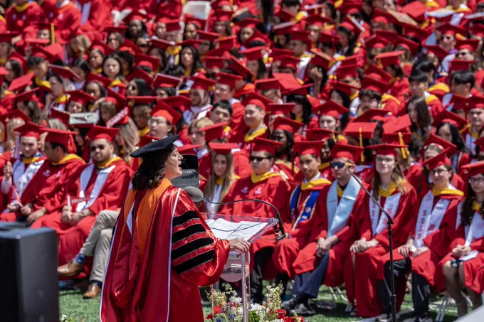 I am speaking at a podium during a graduation ceremony at Stony Brook University. A large group of graduates in red caps and gowns sit in the background on a grassy area.